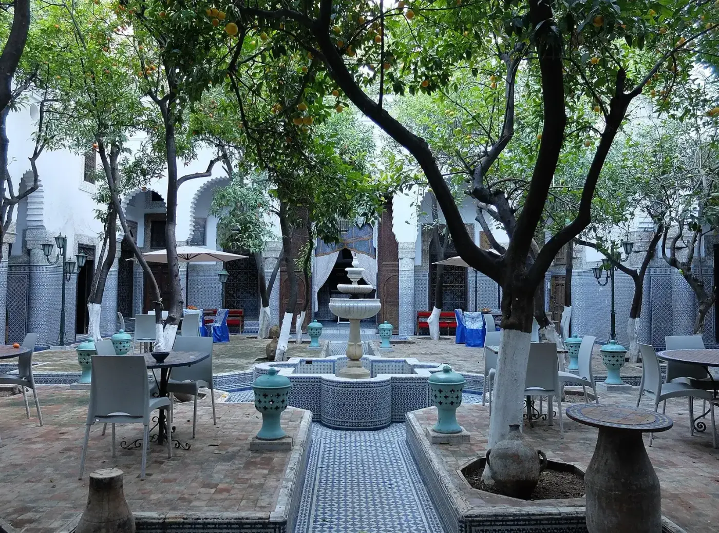 Patio d’une maison de Fès : orangers, fontaine et bassin de zellige au centre, murs carrelés, portes en arc autour de la cour ouverte au ciel.