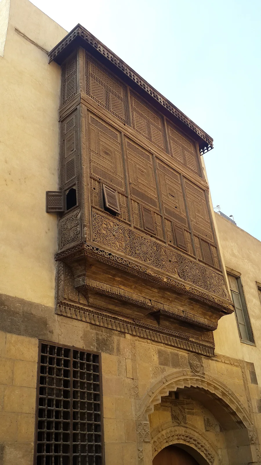 Moucharabieh en saillie sur un mur de pierre au Caire : un grand coffre de bois tourné et sculpté, en encorbellement au-dessus d’un portail.