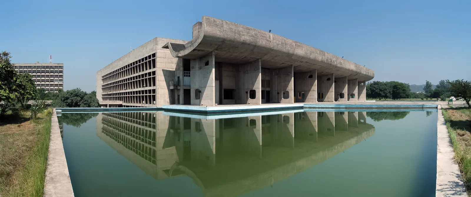 Vue panoramique du Palais de l’Assemblée de Chandigarh : long portique de béton, grand auvent incurvé et tour tronconique, devant un bassin.