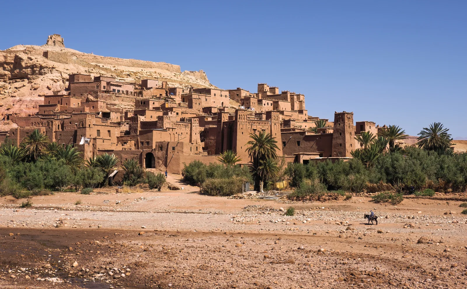 Le ksar d’Aït Ben Haddou vu du lit de la rivière : des maisons de terre serrées sur la pente, des tours, des palmiers au pied du village, un ciel bleu.